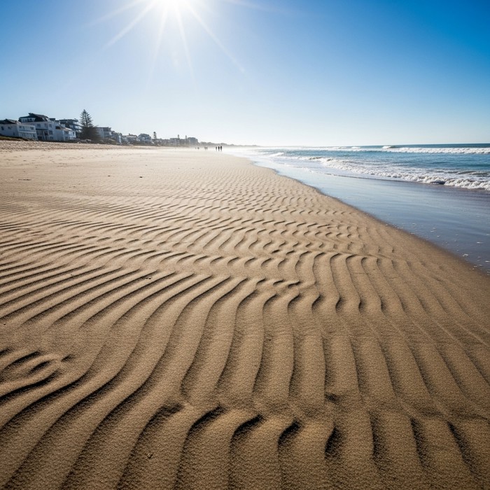 A wide sandy beach with visible ridges of freshly deposited sand left behind by retreating waves, forming delicate ripple patterns under bright sunlight