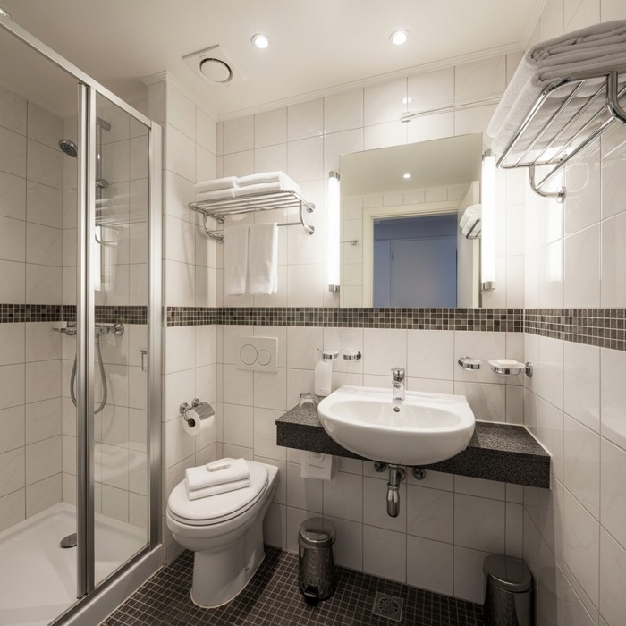A compact hotel bathroom interior spotlessly clean with sparkling tiles, a polished mirror, and bright overhead lighting reflecting off every surface.